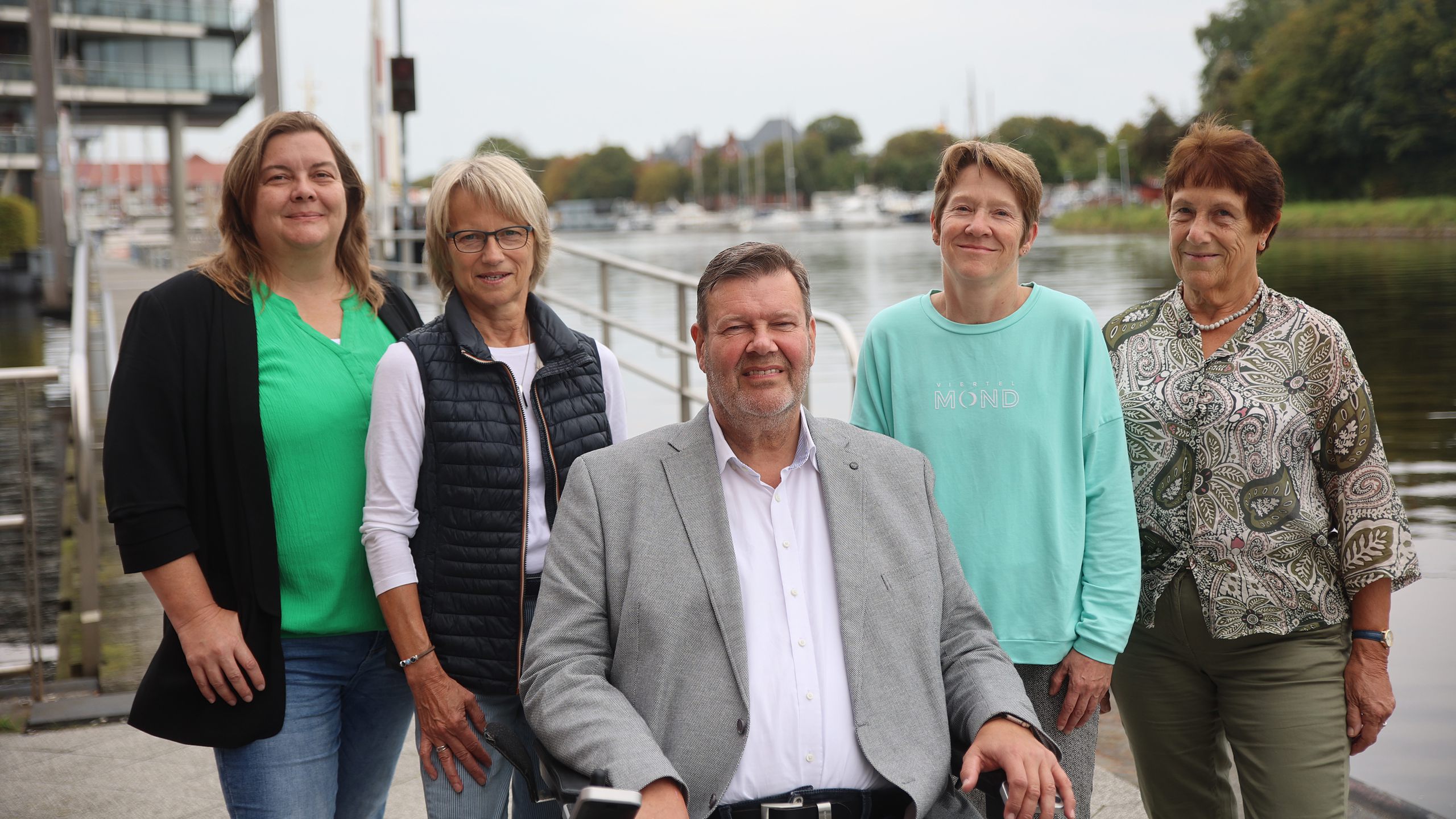 Das Gruppenbild zeigt das Team vom Teilhabebeirat. Vier Personen (von links nach rechts, Sandra Meyer, Anne Groeneweg, Sonja Renken-Muehlbacher, und Elfriede Wilts) stehen am Emder Delft. Ein Mann (Jörn Malanowski) sitzt im Rollstuhl, trägt einen grauen Anzug und ein weißes Hemd. Die Frauen stehen hinter ihm, eine trägt ein grünes Oberteil, eine eine schwarze Weste und die anderen zwei hellgrüne und gemusterte Kleidung.